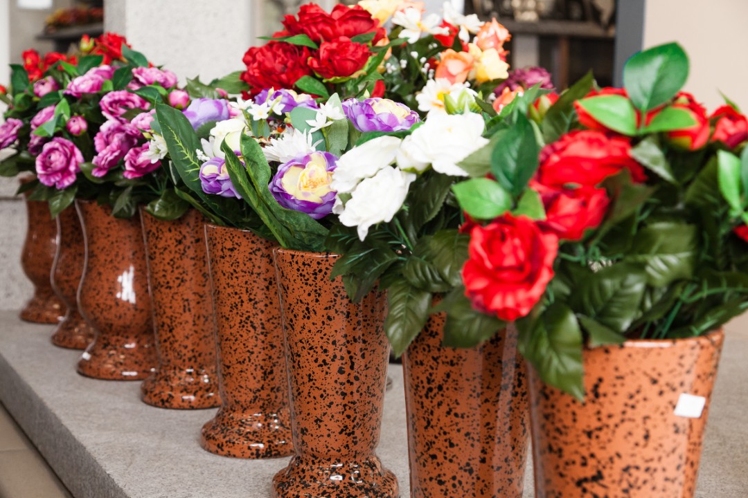 Un alignement de vases en terre cuite brune contenant des fleurs artificielles colorées, y compris des roses rouges, des violettes et des blanches, disposés sur une surface grise dans un environnement intérieur. Ce décor floral apporte une touche de vitalité, illustrant des actualités maison fontan.